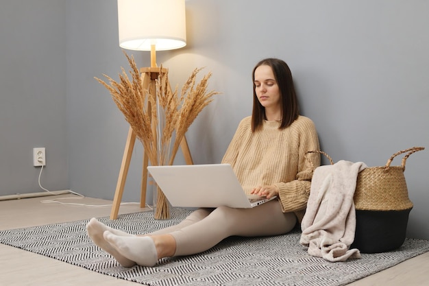 Freelancers workspace Remote job opportunities Concentrated calm brown haired woman wearing beige sweater working on computer while sitting on floor in home interior looking at display