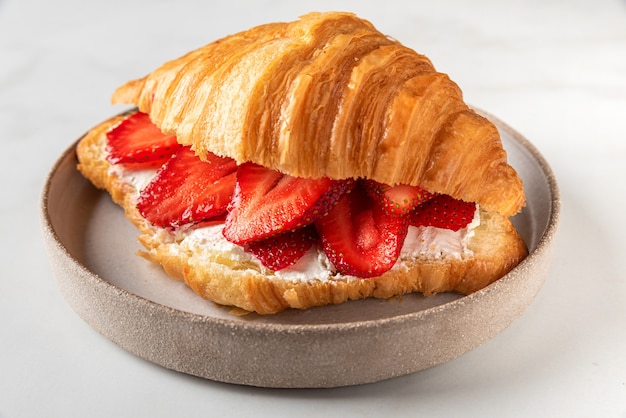 French croissant sandwich with fresh ripe strawberries and cream cheese in a plate on white marble surface