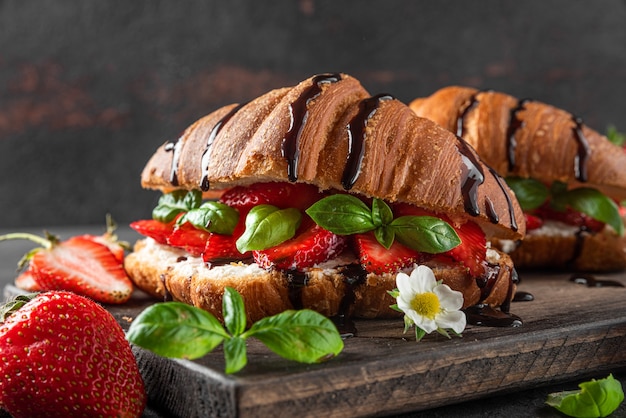 Photo french croissant sandwiches with fresh strawberries, cream cheese, basil and chocolate sauce on dark surface