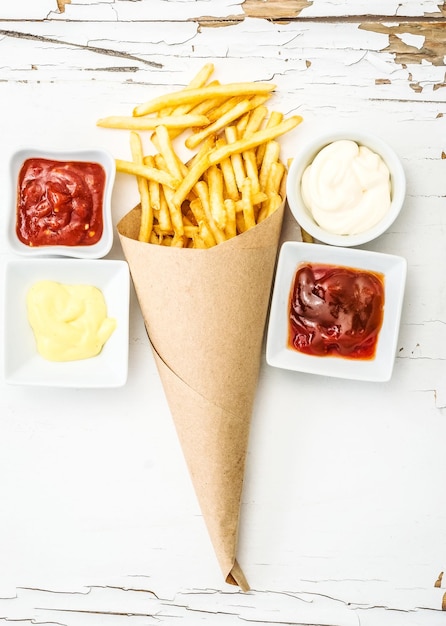 French fries wrapped in paper among different sauces in white saucers on the white wooden background