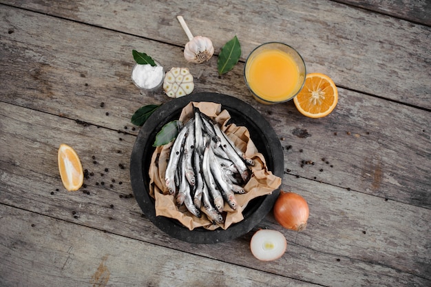 Fresh anchovies on plate on the baking paper with fresh orange juice, garlic, onion and lemon around on the wooden table