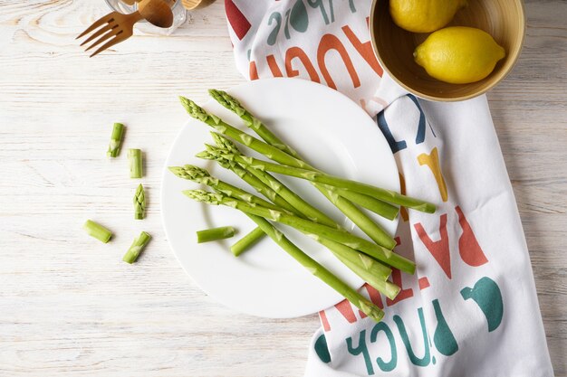 Fresh asparagus on a plate and lemons on a light wood surface. Top view.