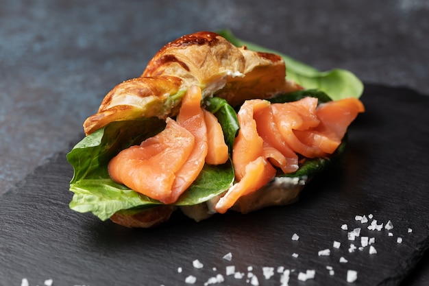 Fresh baked croissant sandwich with salad and salmon on a black slate board