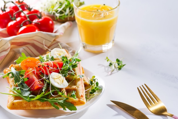 Fresh baked sweet belgian waffles with eggs, tomatoes, micro green and avocado served on a plate with orange juice on marble 