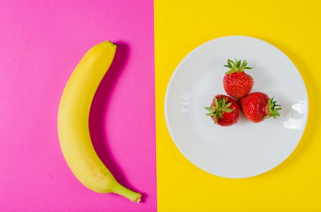 A fresh banana and strawberries lie on a yellow and pink surface