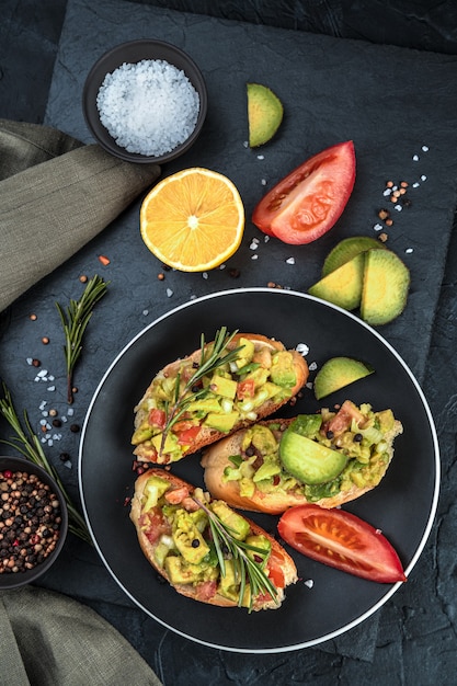 A fresh breakfast of sandwiches with avocado, tomatoes, lemon and rosemary on a black background. Top view, vertical orientation. The concept of healthy eating.