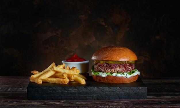 Fresh burger with french fries on dark wooden board and bowl of tomato sauce