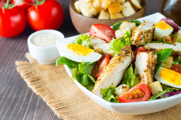 Fresh Caesar salad with delicious chicken breast, ruccola, spinach, cabbage, arugula, egg, parmesan and cherry tomato on wooden background. Oil, salt and pepper. Healthy and diet food concept.