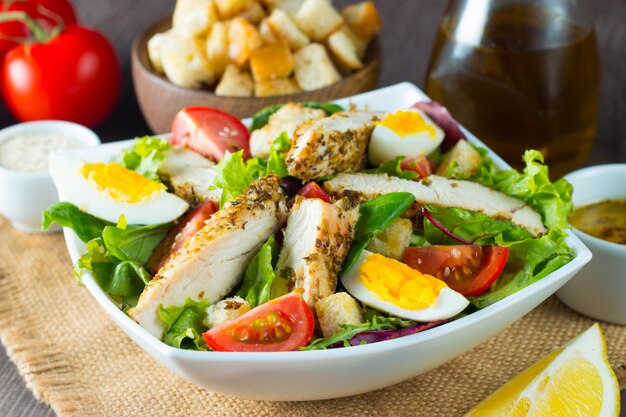 Fresh Caesar salad with delicious chicken breast, ruccola, spinach, cabbage, arugula, egg, parmesan and cherry tomato on wooden background. Oil, salt and pepper. Healthy and diet food concept.