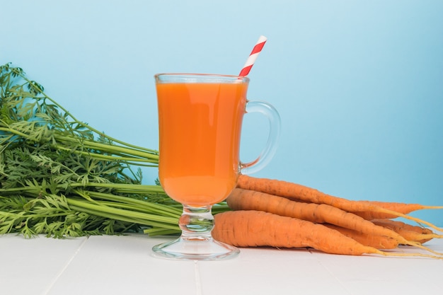 Fresh carrots and a glass with a smoothie and a cocktail tube on a blue background.