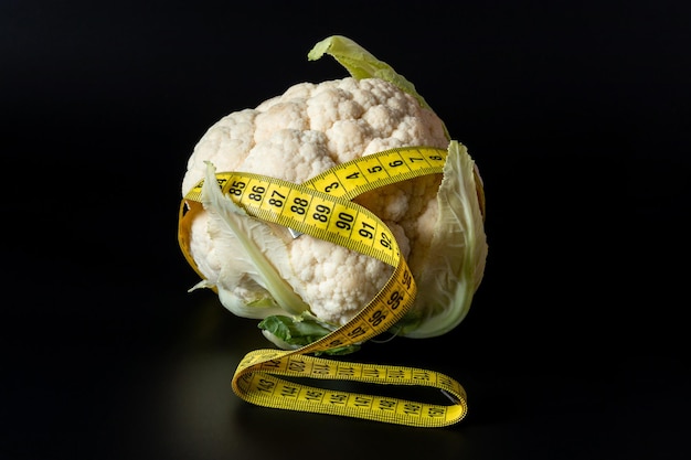 A fresh cauliflower head wrapped with a measuring tape on a black background. The concept of healthy nutrition, weight loss, dietetics