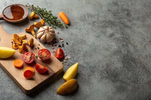 Fresh cherry tomatoes, potatoes, mushrooms chanterelles, carrot, herbs on cutting board and wooden cooking spoon with olive oil. Food cooking background with ingredients, top view, space for text