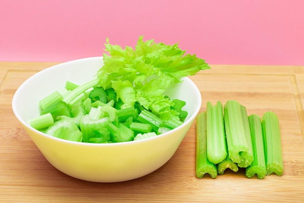 Fresh chopped celery slices in white bowl with celery sticks on bamboo cutting board