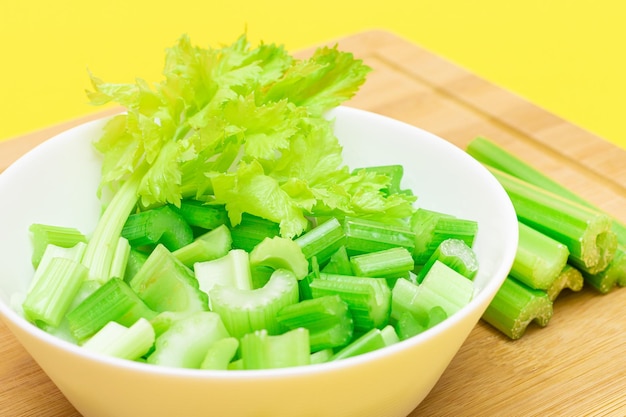 Fresh chopped celery slices in white bowl with celery sticks on bamboo cutting board