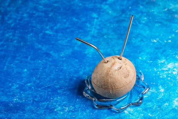 Photo fresh coconut with two metal straw inserted in it on glass plate on blue background