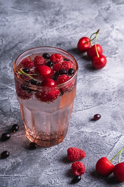 Fresh cold sparkling water drink with cherry, raspberry and currant berries in red faceted glass on stone concrete background, summer diet beverage, angle view