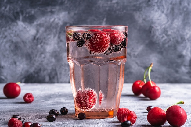 Fresh cold sparkling water drink with cherry, raspberry and currant berries in red faceted glass on stone concrete surface, summer diet beverage, angle view