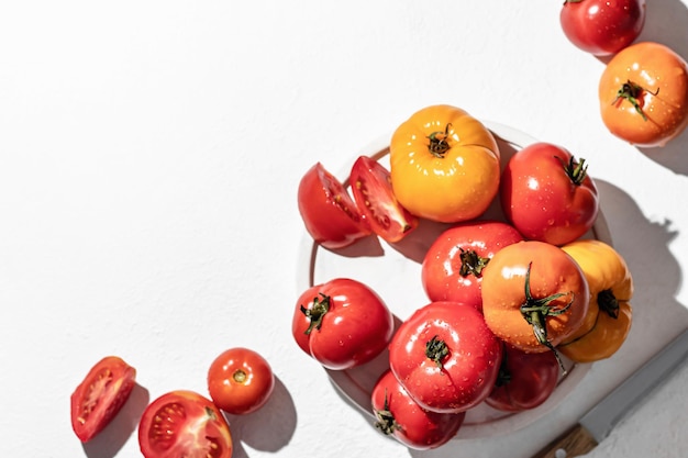 Fresh colorful tomatoes and knife on plate with hard shadows on white with copy space top view