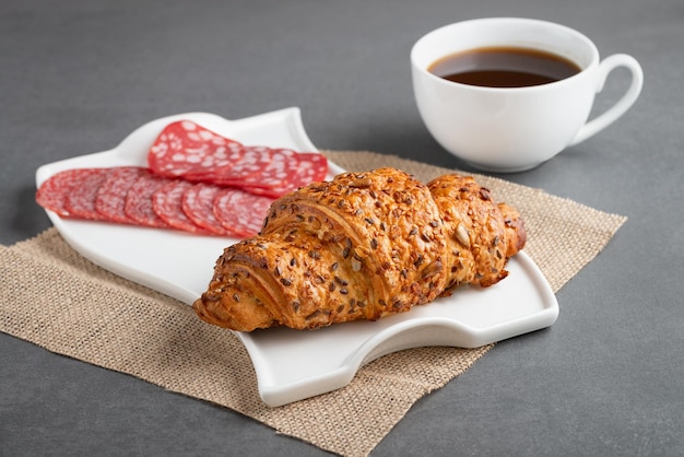 Fresh croissant sandwich with sausage and coffee on grey table