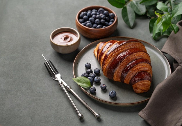 Fresh croissant with chocolate and blueberries on a dark plate on a green background with branch Delicious homemade breakfast concept in rustic style