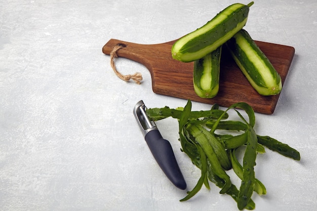 Fresh cucumbers with skin and knife on a brown wooden cutting board on a light gray concrete table