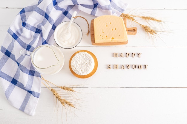 Fresh dairy products apples a bouquet of ears of corn on a white wooden background with wooden letterstext of happy shavuot top view