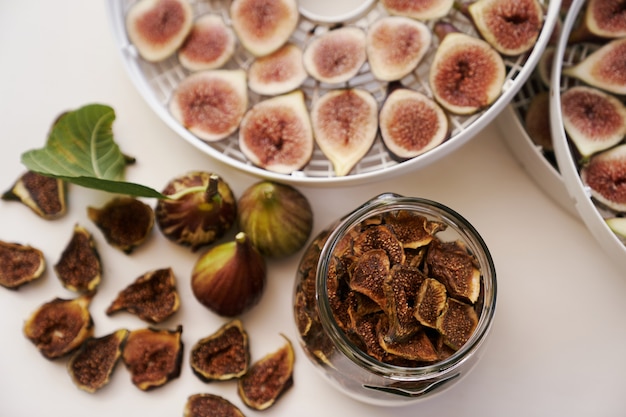 Fresh figs lie on the table next to dried figs in a glass jar and a dehydrator top view