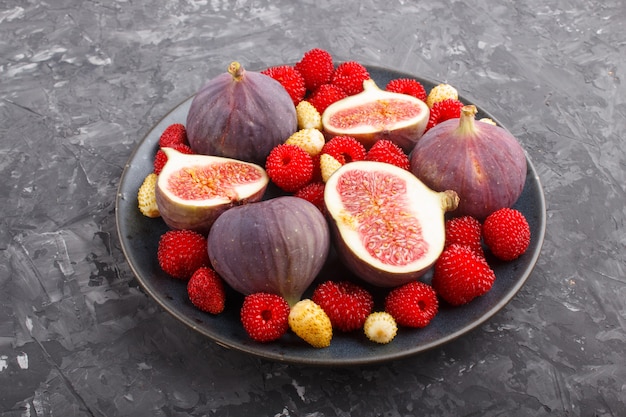 Fresh figs, strawberries and raspberries on blue plate on black concrete surface. side view, close up.