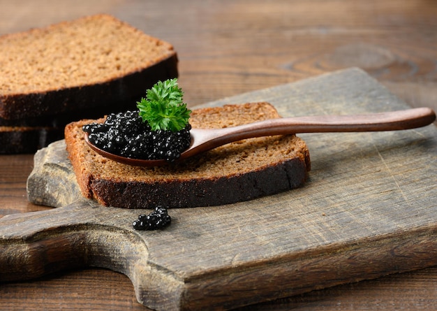 Fresh grainy black paddlefish caviar in brown wooden spoon on a brown table, close up