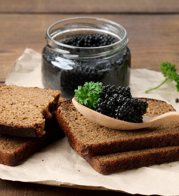 Fresh grainy black paddlefish caviar in brown wooden spoon on a brown table, close up