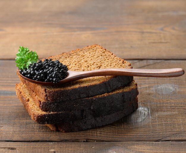 Fresh grainy black paddlefish caviar and slices of bread in brown wooden spoon on a brown table, close up