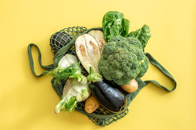 Fresh green vegetables in a green string bag on a yellow background flat lay