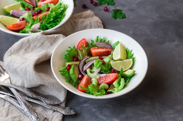 Fresh healthy salad with tomatoes, avocado and pomegranate in bowls on gray concrete.