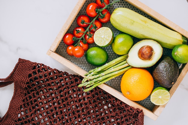 Fresh healthy vegetables in wooden box and eco shopping bag on white marble background