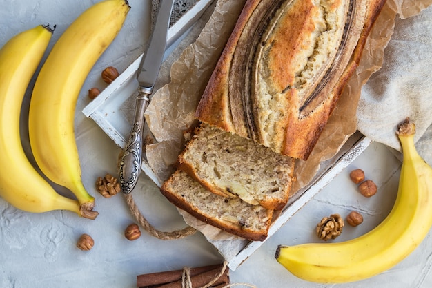 Fresh homemade banana bread in white wooden tray with ingredients on light concrete surface. Top view.