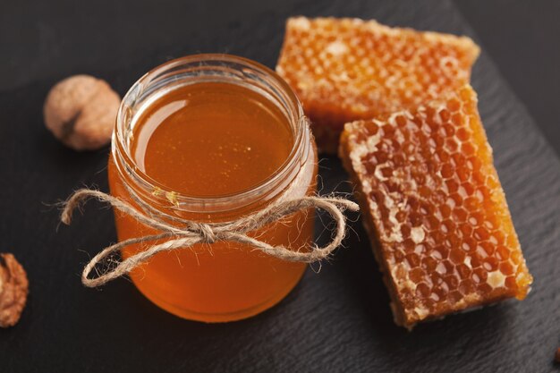 Fresh honeycombs and liquid honey in glass jar on black background, closeup