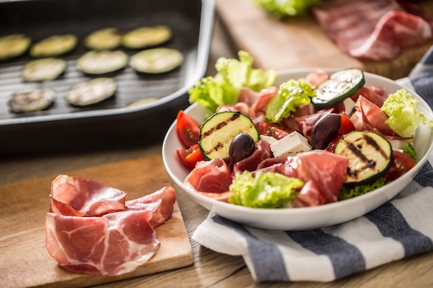 Fresh lettuce salad with grilled zucchini coppa di parma ham feta cheese olives tomatoes and olive oil.