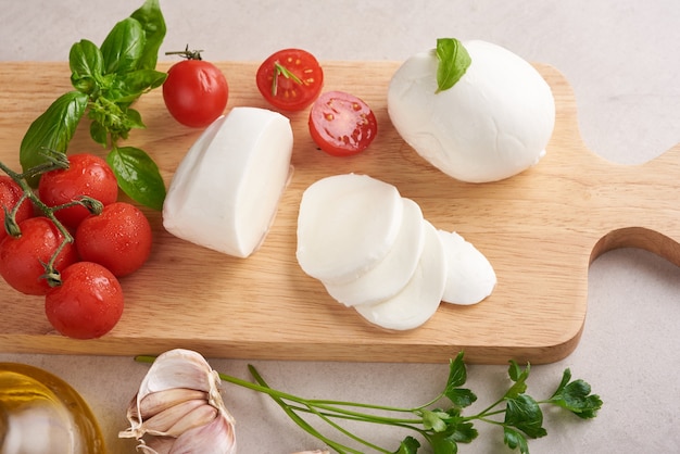 Fresh mozzarella cheese, Soft italian cheeses, tomato and basil, olives oil and rosemary on wooden serving board over light wooden surface. Healthy food. Top view. Flat lay.