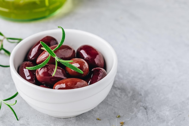 Fresh organic black olives in olive oil in white bowl with rosemary on gray stone background