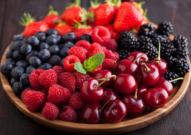 Fresh organic summer berries mix in round wooden tray on dark wooden table background. Raspberries, strawberries, blueberries, blackberries and cherries. Top view