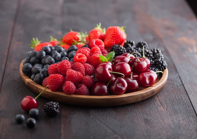 Fresh organic summer berries mix in round wooden tray with glass of juice on dark wooden table background. Raspberries, strawberries, blueberries, blackberries and cherries.  Top view