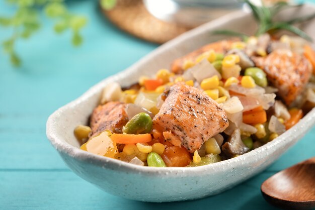 Fresh pan-fried salmon cube chops with stir-fried mixed vegetables including edamame beans, carrot, onion, mushroom and corn kernels on blue wooden table surface.