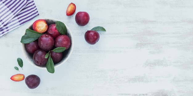 Fresh plums with leaves in bowl on white rustic wooden table background. Autumn harvest concept. Top view with copy space for text