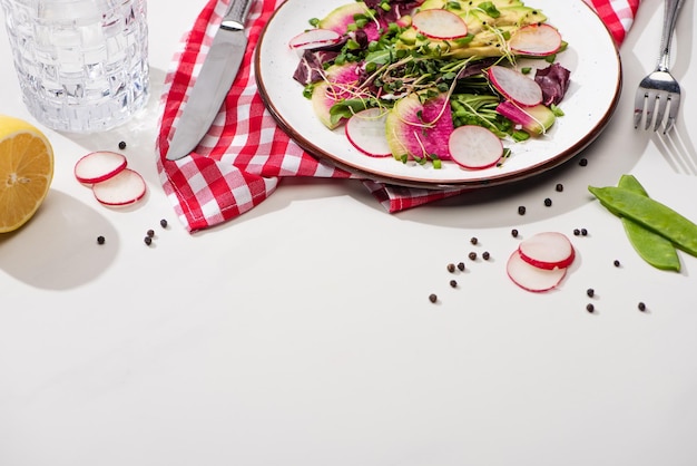 Fresh radish salad with greens and avocado on plate on white surface with water and cutlery