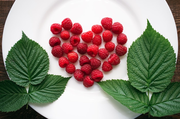 Fresh raspberry in heart shape with leaves on white plate