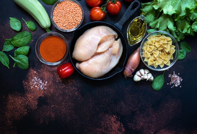 Fresh raw chicken meat, chicken fillet in a pan surrounded by healthy food ingredients on a dark rustic background. Top view, copy space