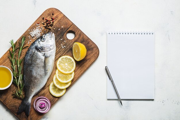 Photo fresh raw dorado fish on a rustic wooden cutting board with spices and a notebook for recipe or menu. top view on a white background.