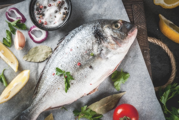 Fresh raw organic fish Dorado, sea bream with ingredients for cooking lemon, tomatoes, onion, salt, pepper, bay leaf, greens, oil. On old wooden cutting board, on black stone table Top view copy space