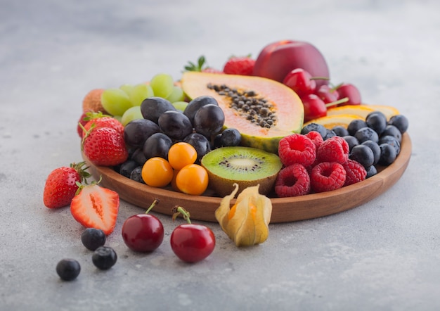 Fresh raw organic summer berries and exotic fruits in round wooden plate on light kitchen background. Papaya, grapes, nectarine, orange, raspberry, kiwi, strawberry, lychees, cherry. Top view