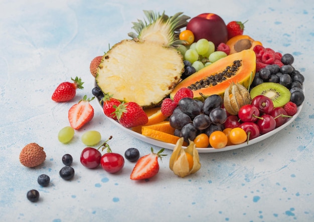 Fresh raw organic summer berries and exotic fruits in white plate on blue kitchen background Pineapple papaya grapes nectarine orange apricot kiwi pear lychees cherry and physalis Top view